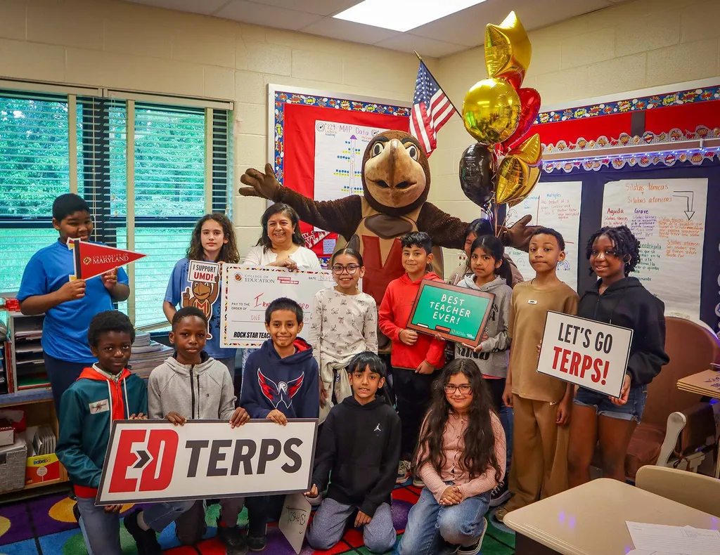 teacher holds giant check and poses with students and Testudo in classroom. Students hold UMD signs and balloons