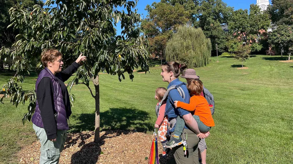 a woman points at a tree in a field while a woman carrying two children looks on