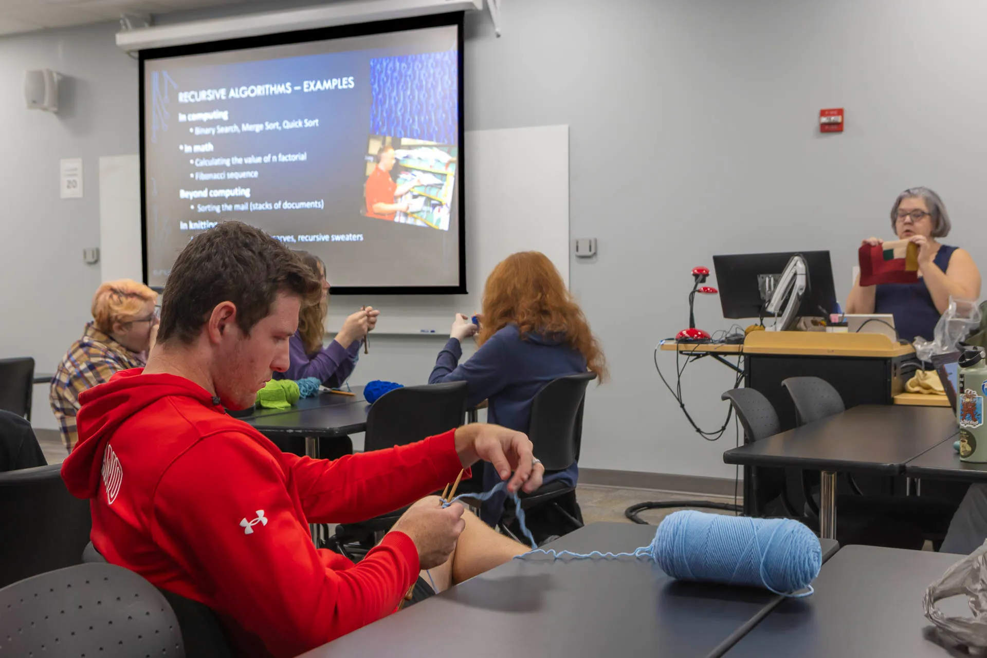student in red sweatshirt knits with blue yarn, with slide that reads, "recursive algorithms - examples" in the background