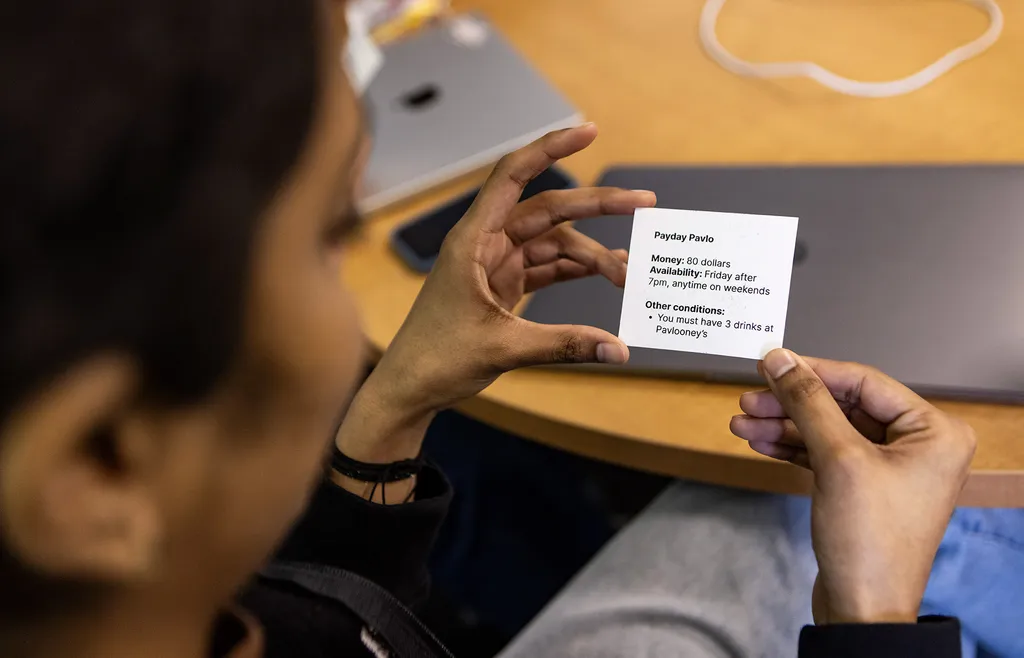 student holds conditions card: "Payday Pavlo. Money: $80. Availability: Friday after 7 p.m., anytime on weekends. Other conditions: You must have 3 drinks at Pavlooney's"