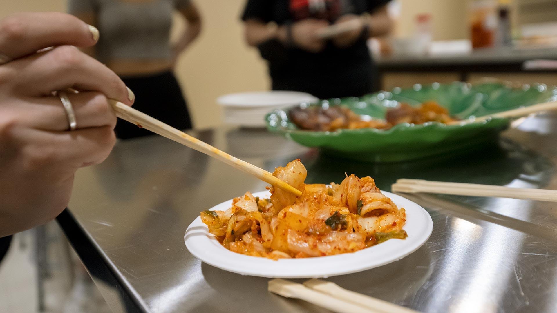 A hand uses chopsticks to grab kimchi from a white plate