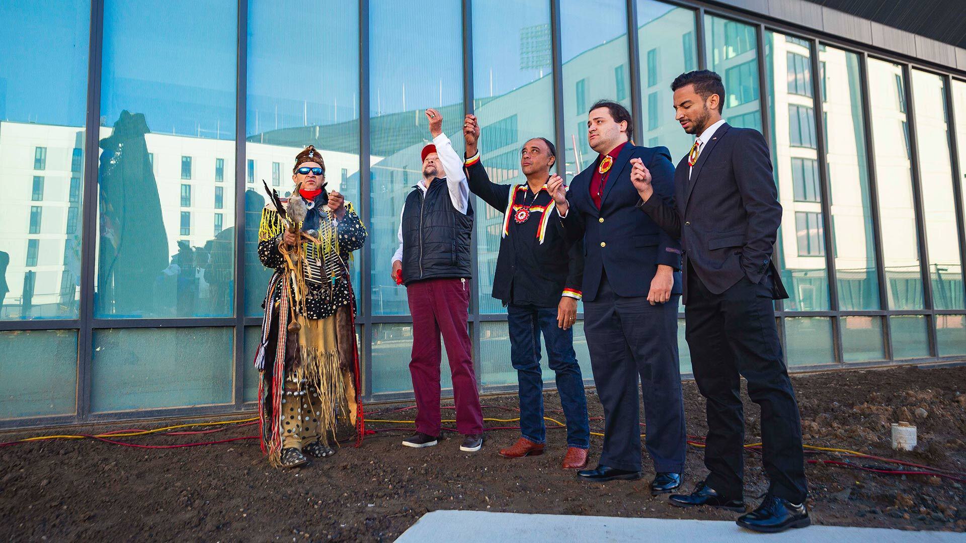 Five men stand in front of reflective windows on dirt, with two raising a hand in the air