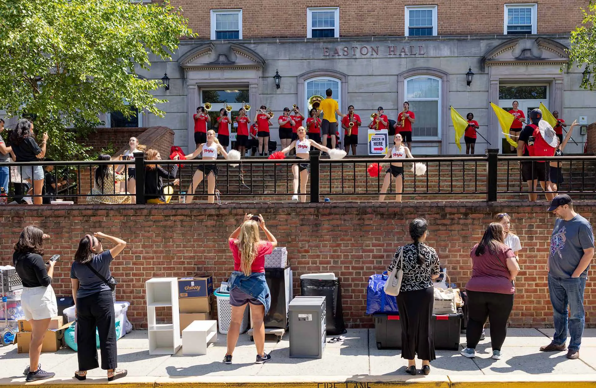 cheerleaders and marching band perform on top of wall outside a residence hall as parents and students observe