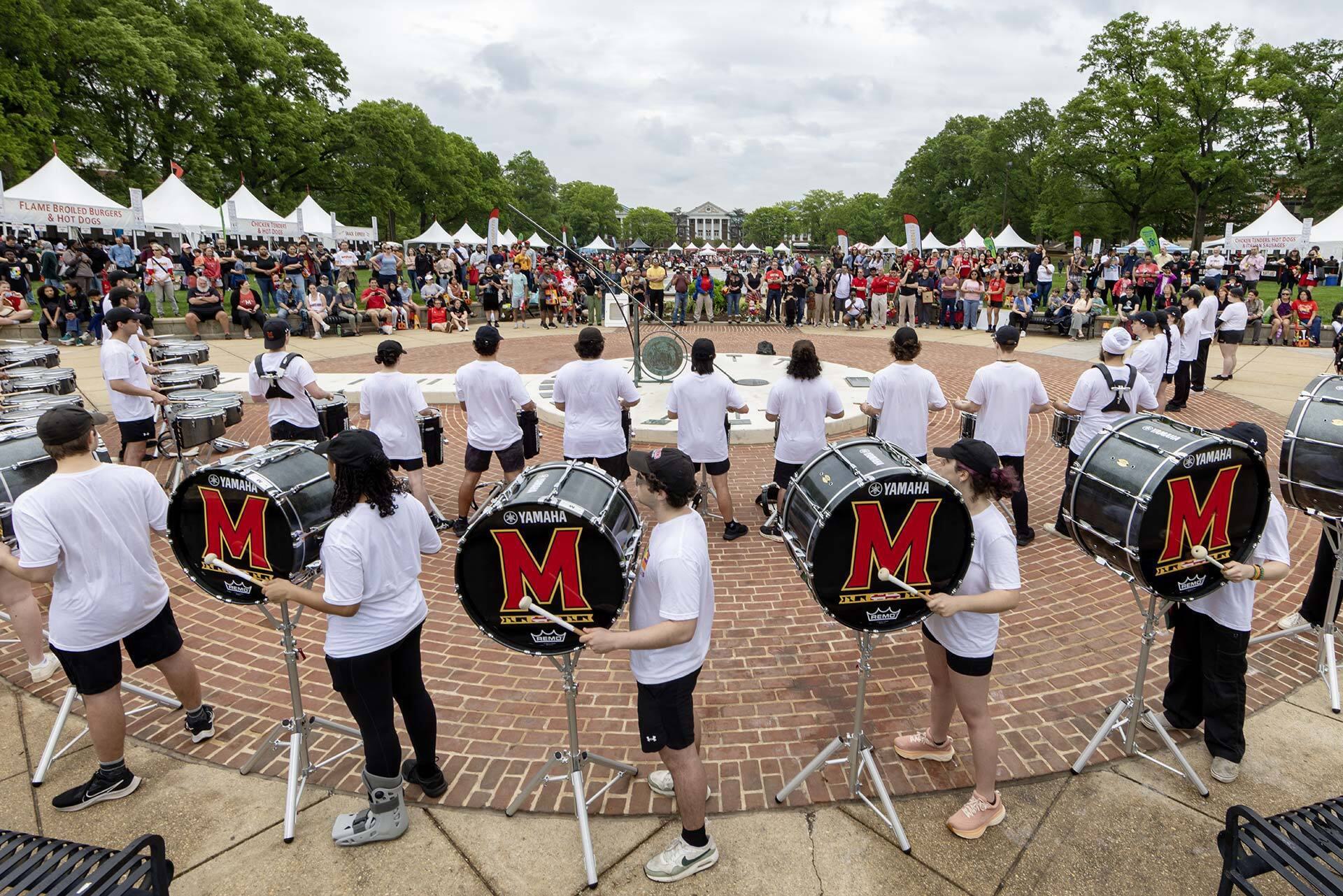 A bunch of drummers outdoors play for a crowd