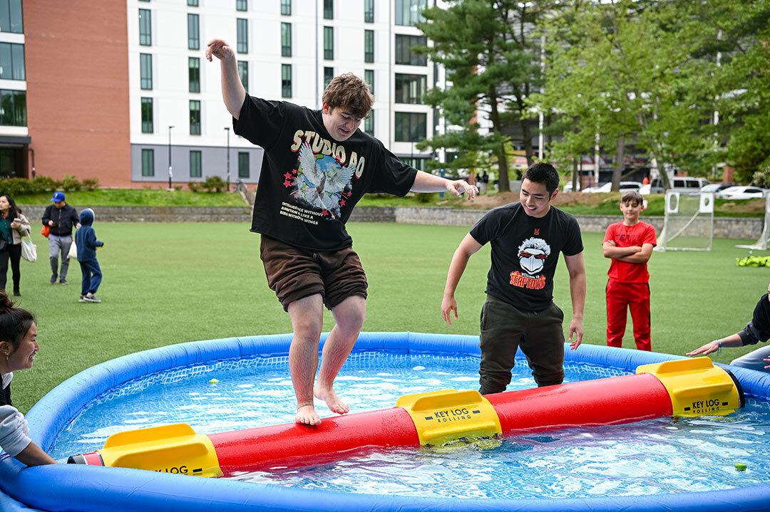 two young men compete in a log rolling competition, and one has fallen into the water