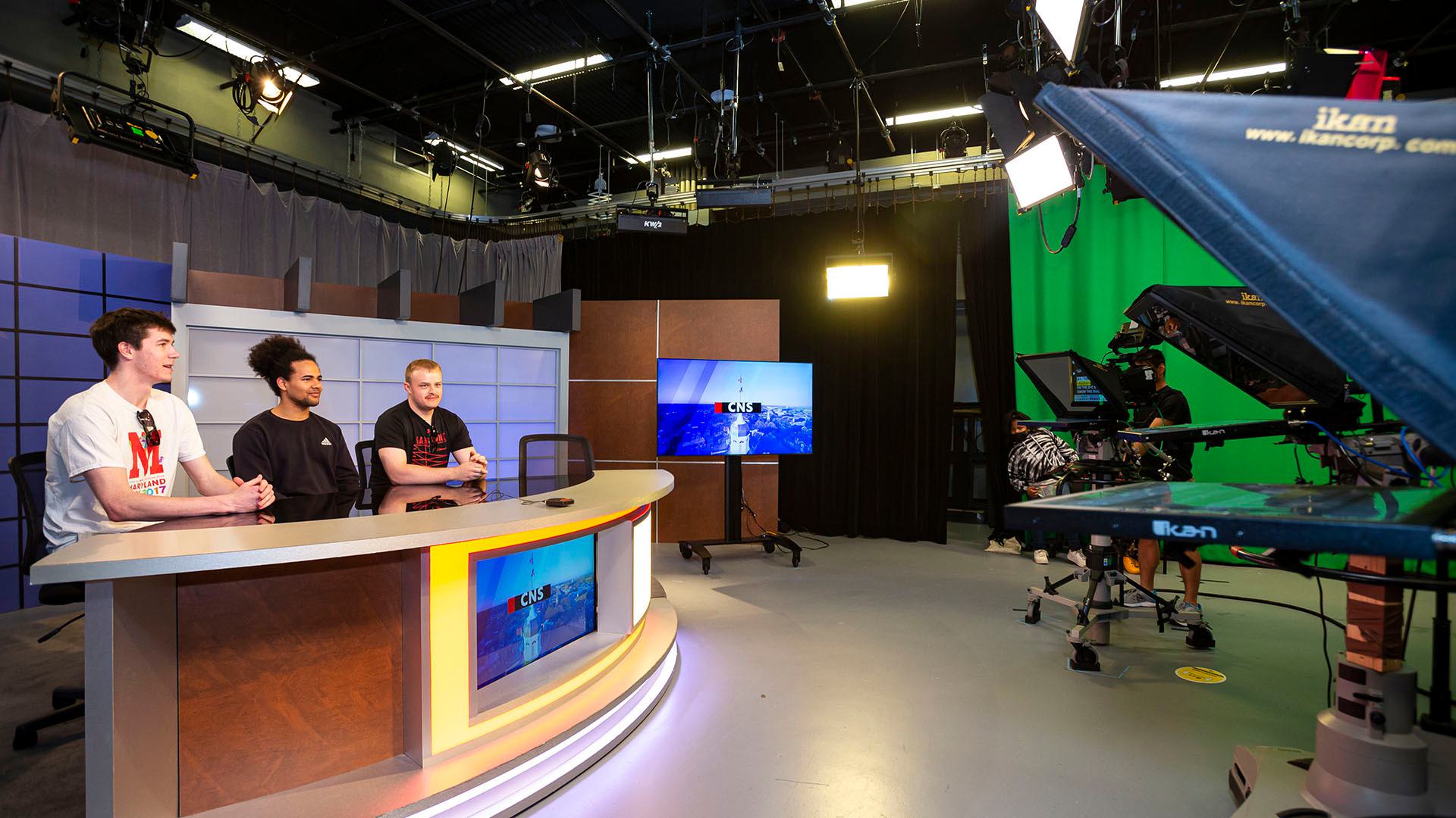Three people sit at news anchor desk