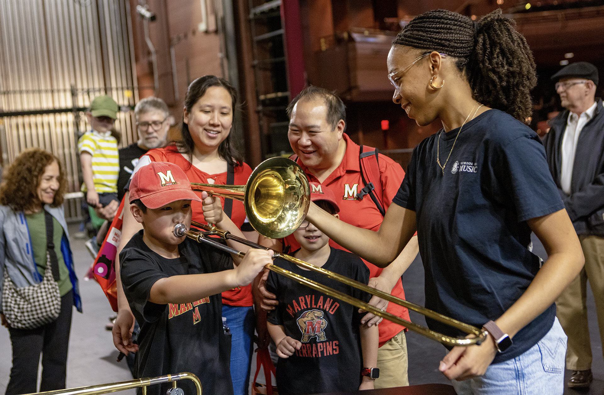 Woman helps young boy play trombone, watched by boy's smiling family