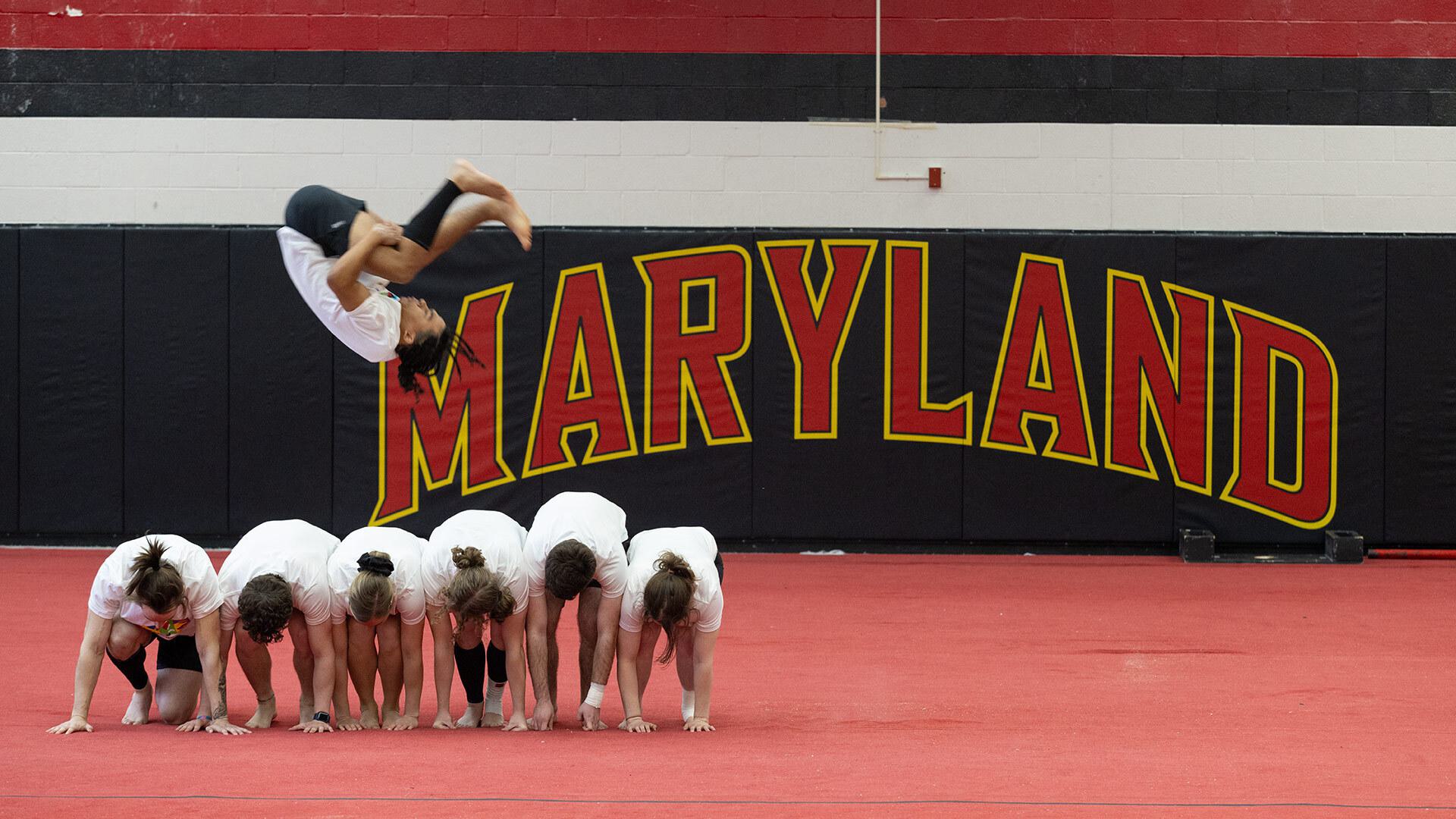 one person does a running flip over five other people who are bending over in front of a Maryland logo on a wall.