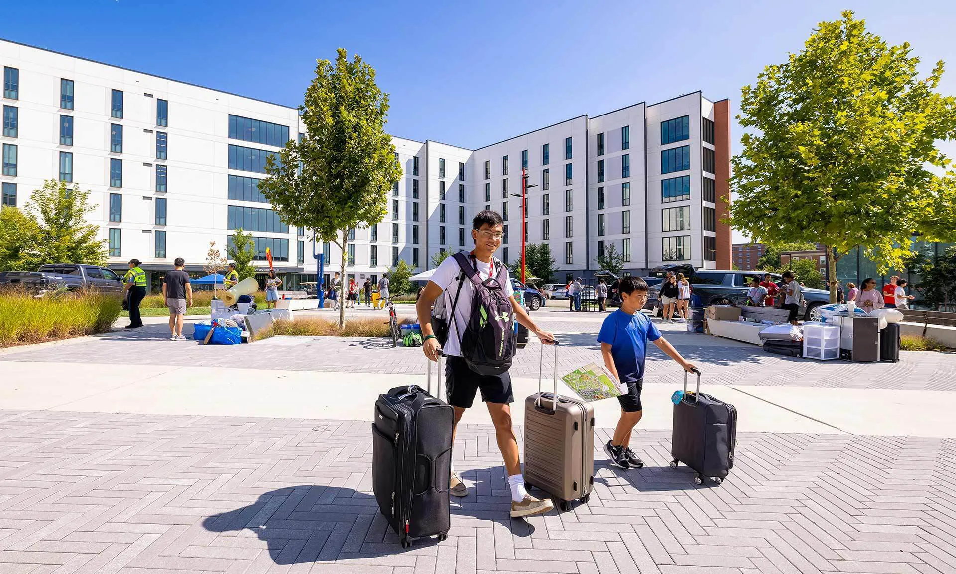 A freshman and his younger brother roll suitcases toward a residence hall on a sunny day