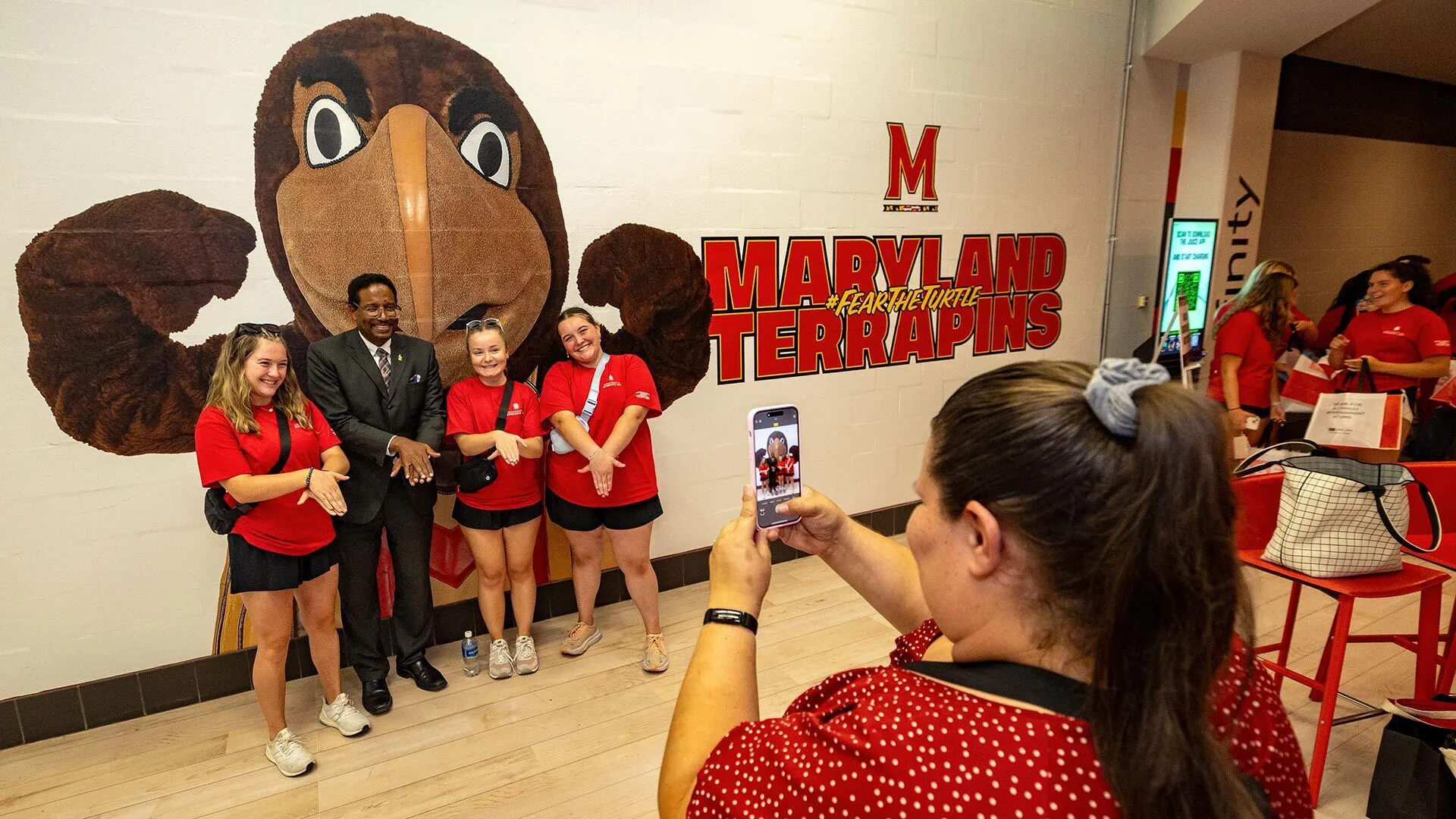 three young women in red t shirts and black shorts pose with middle aged man in dark suit in front of giant mascot mural