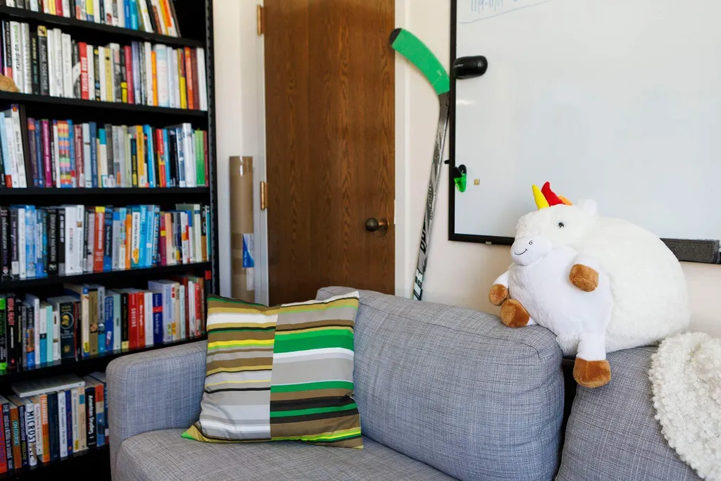 Bookshelf and gray couch with a white stuffed unicorn and a green hockey stick poking out behind it