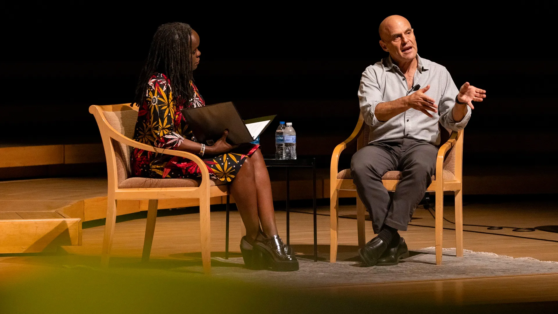 Two people seated in chairs speak on a stage. A woman holds a binder, while the man again gestures.