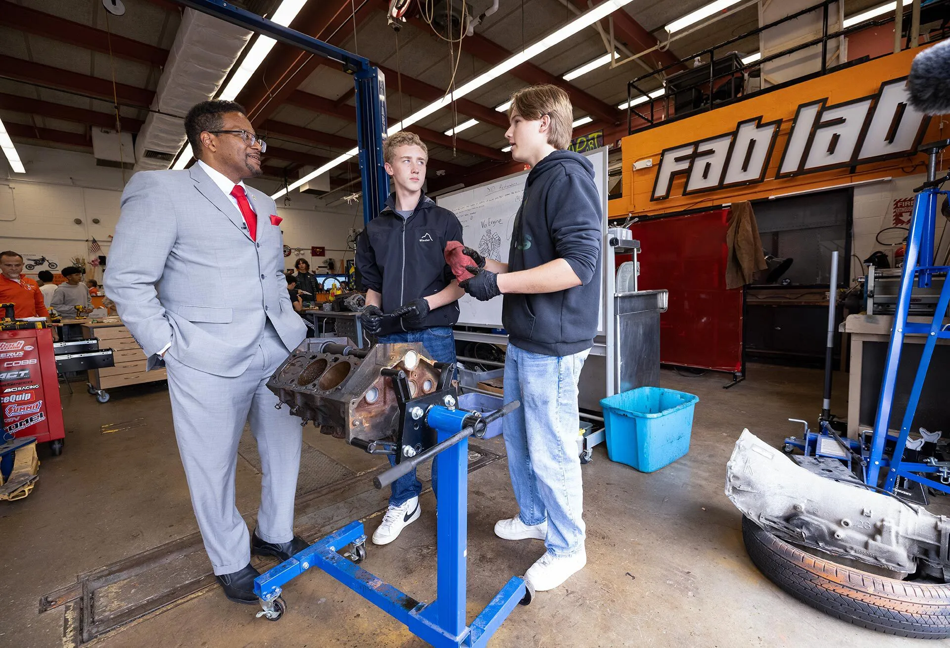 A man in a suit talks to high school students standing near an engine block on a stand.