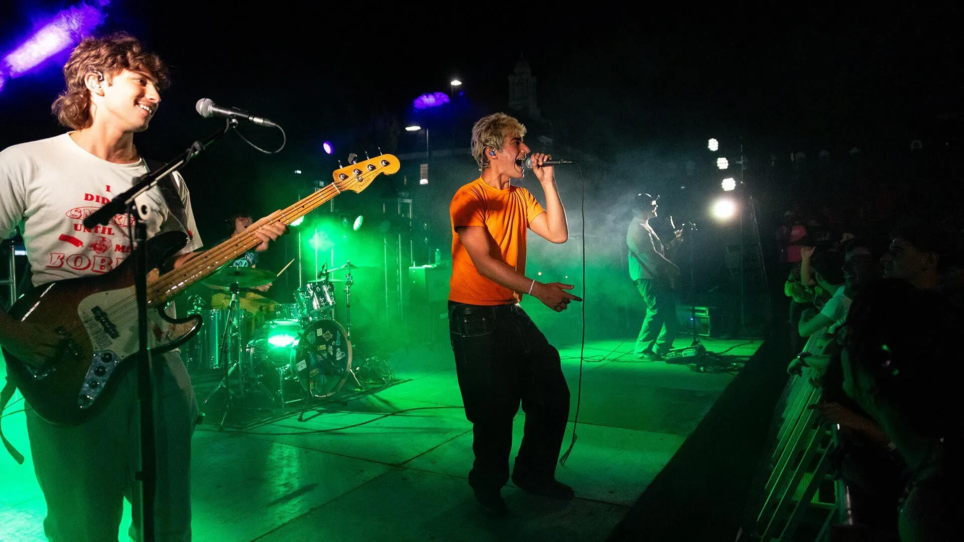 a rock band including a bassist and a lead singer in a birght orange shirtplays onstage under green lighting