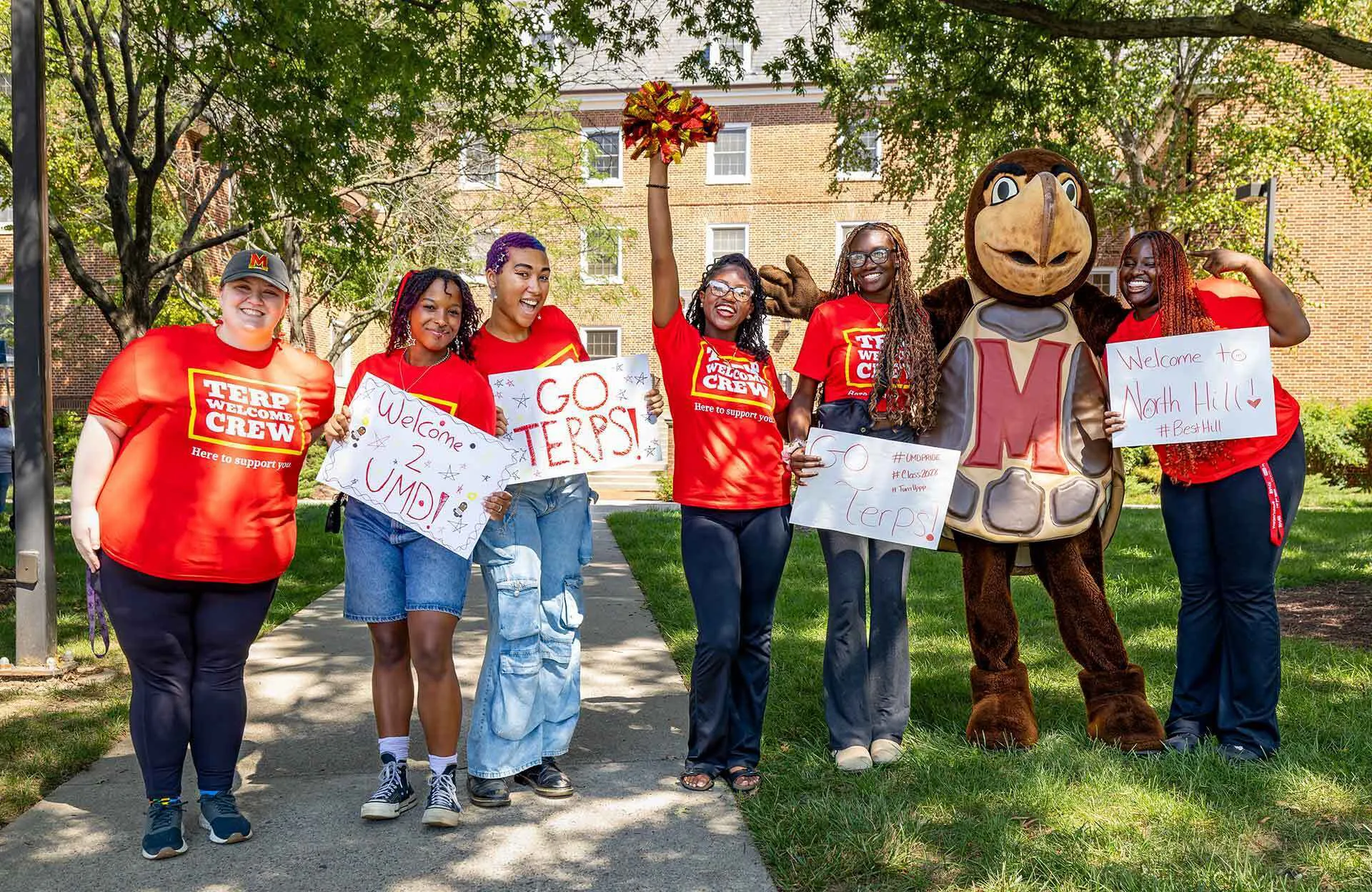 six students holding welcome signs pose with mascot outside a residence hall