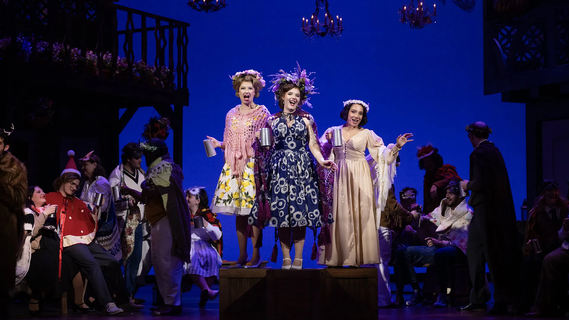 three cast members hold silver mugs and wear flower crowns on stage