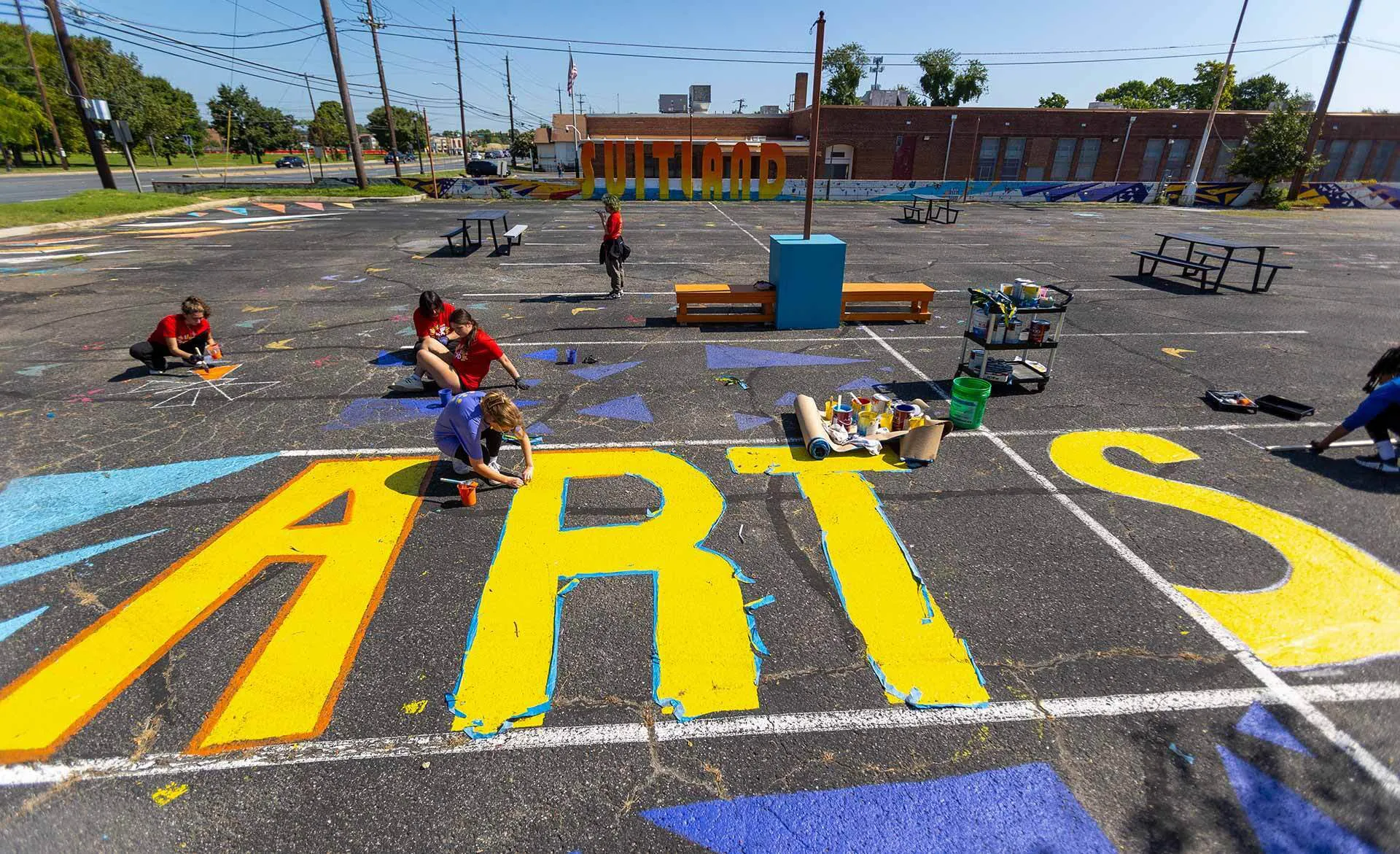 several students sit in a big empty parking lot painting a color mural on ground