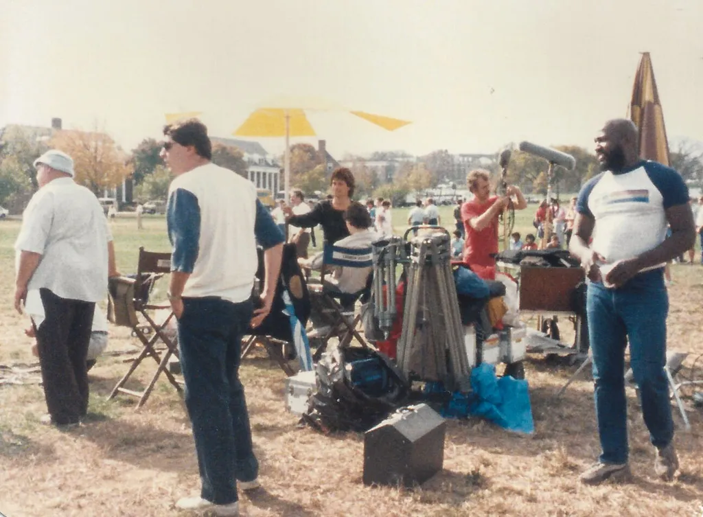 Rob Lowe under a white and yellow striped umbrella in the middle of film equipment and crew on Fraternity Row