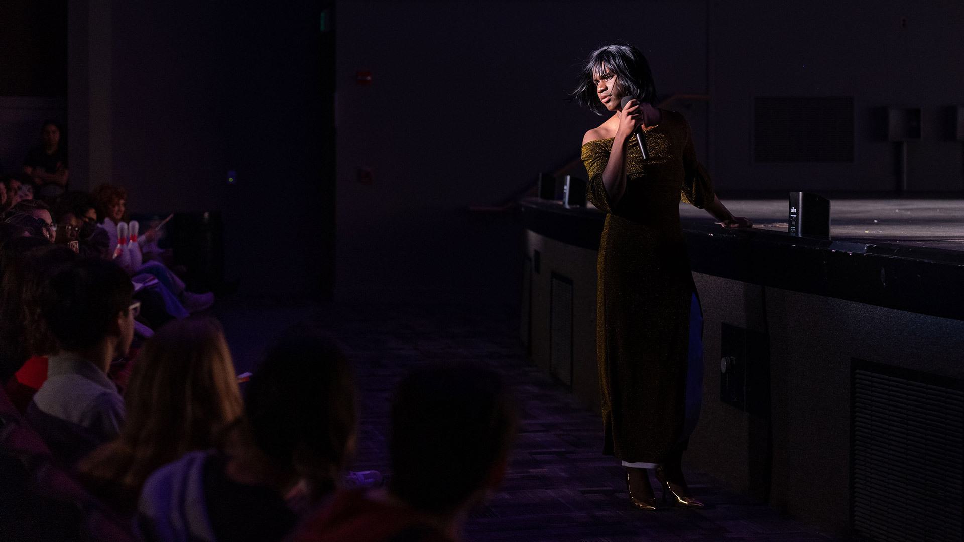Person in a fancy dress holds a microphone in front of a crowd in a dark auditorium