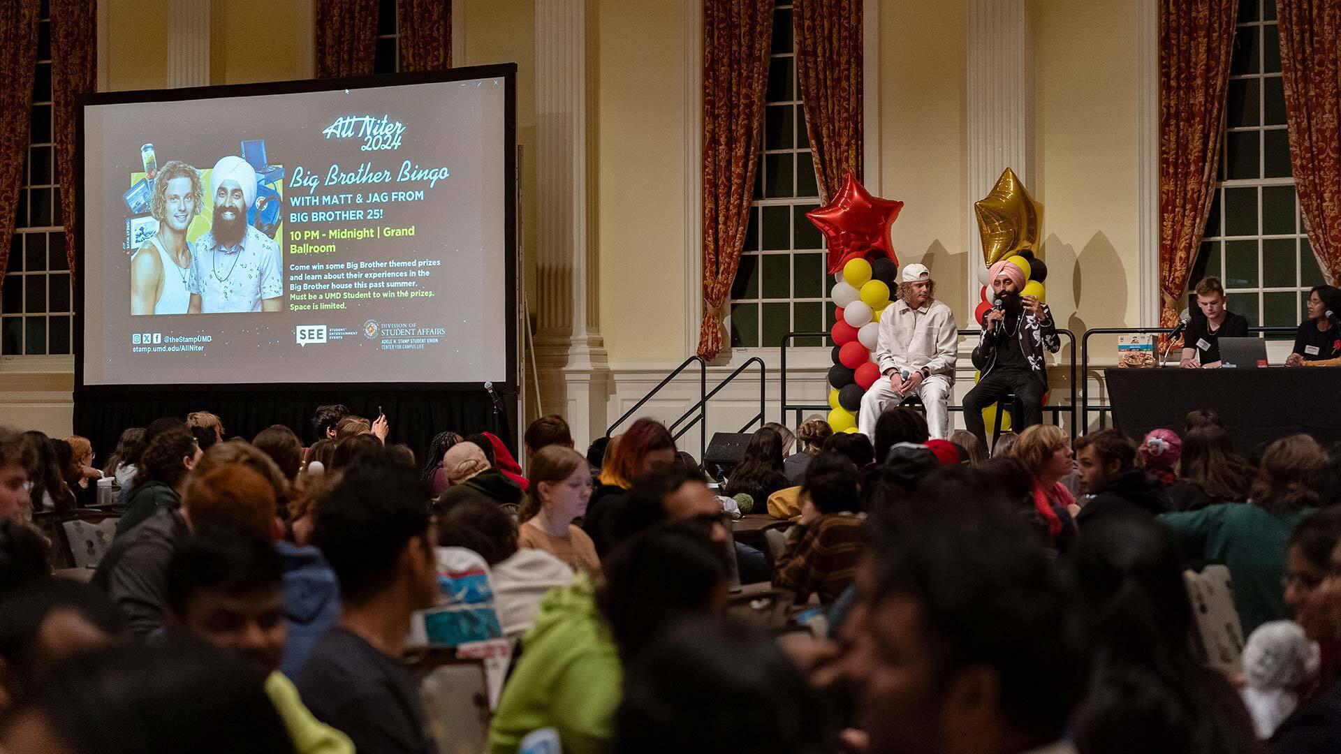 Leaders of a large bingo game sit on a stage next a big screen