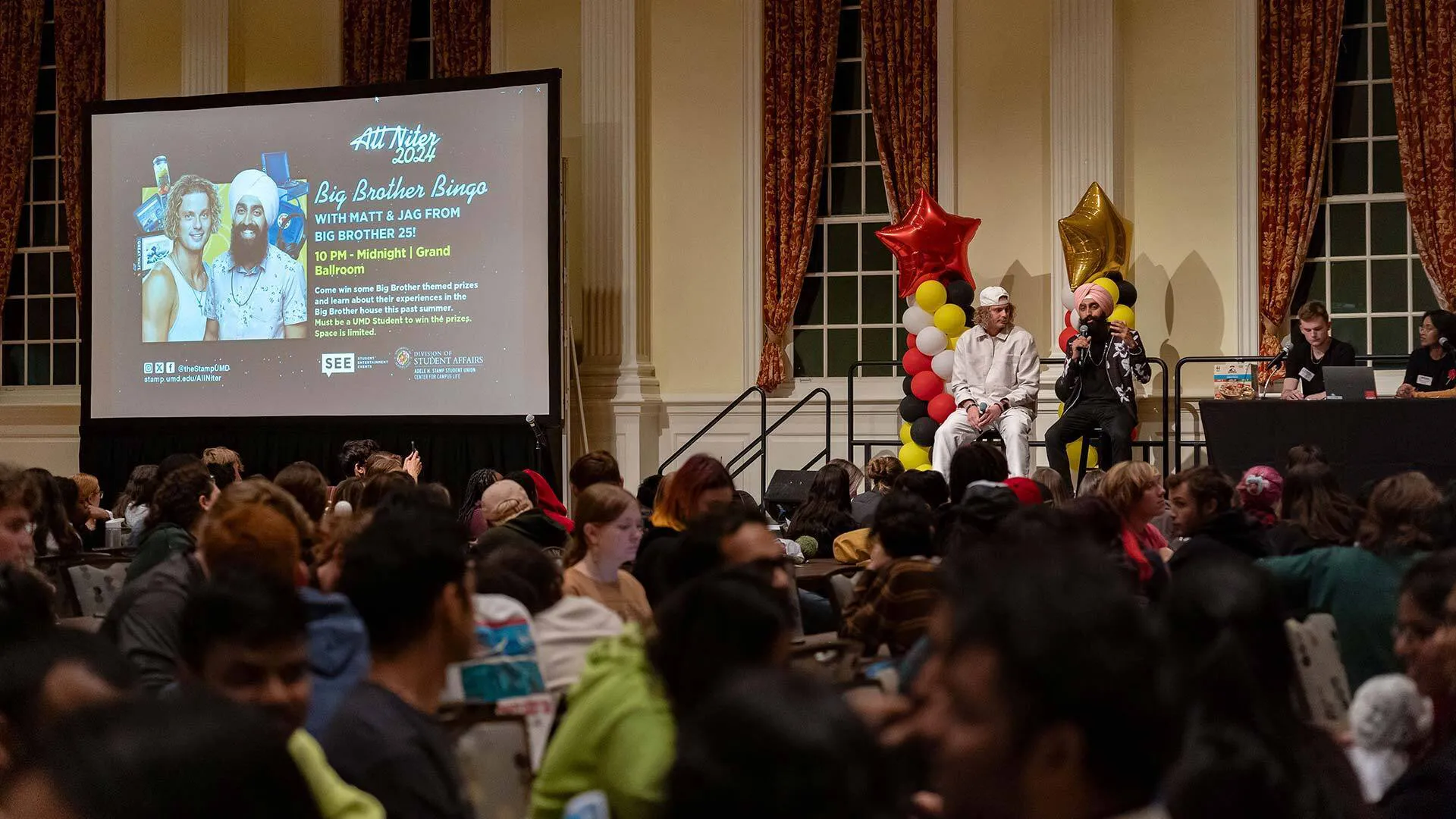Leaders of a large bingo game sit on a stage next a big screen