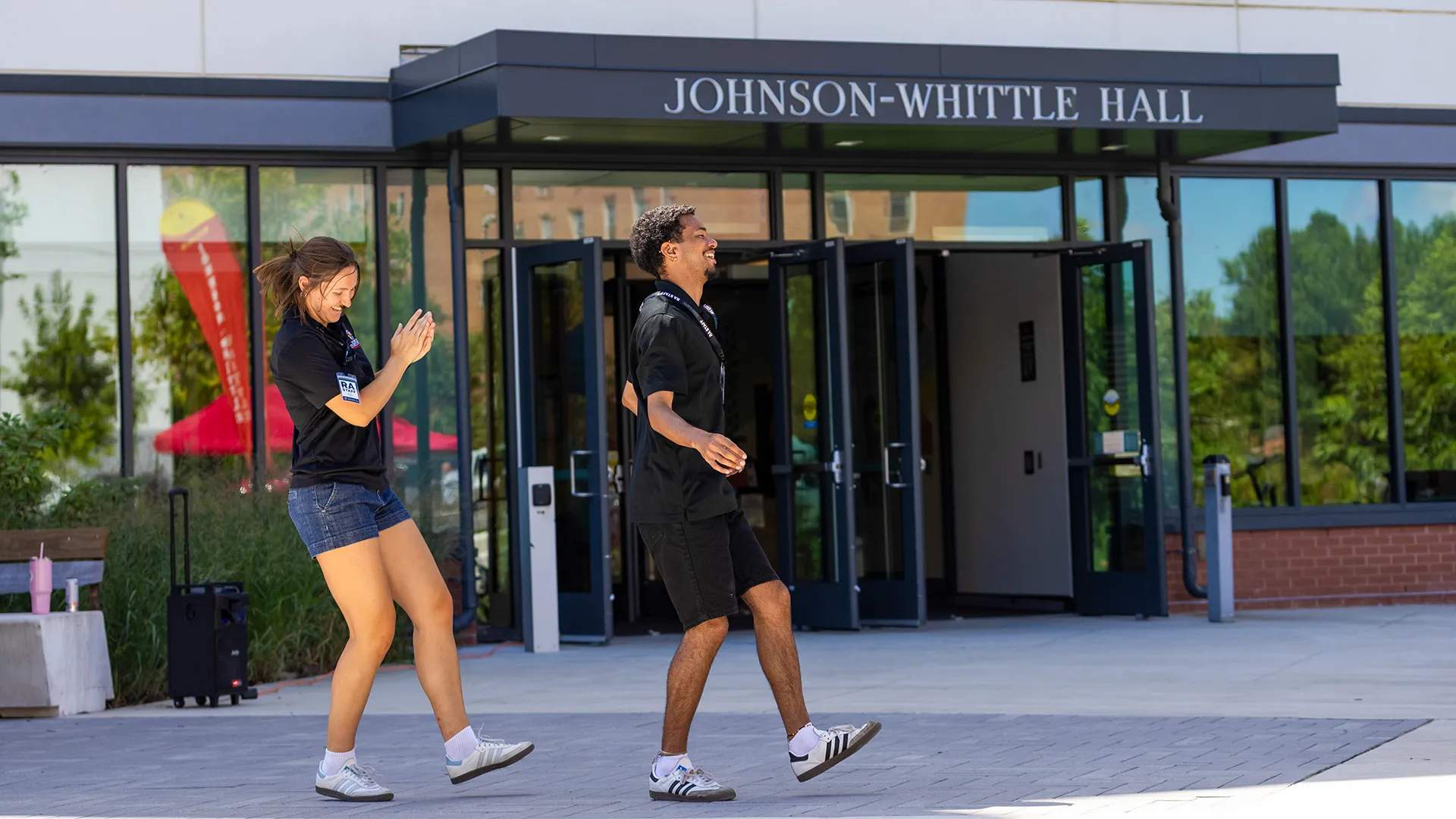 Two people in shorts and black polo shirts dance outside a residence hall entrance.