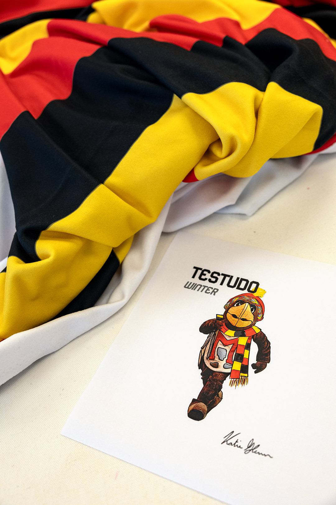 sketch of Testudo in winter clothing, next to red, black and gold fabric