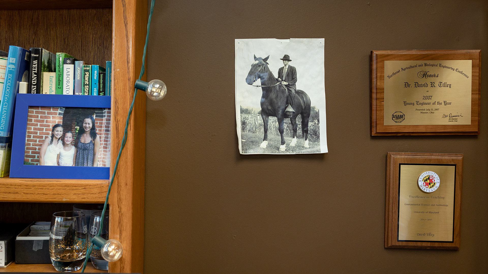 black and white photo of a man in a hat sitting on top of a horse taped to a brown wall, surrounded by awards