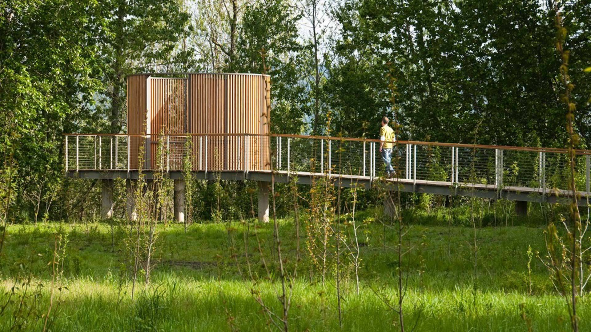 man walks on bridge toward elevated cylindrical blind in woods