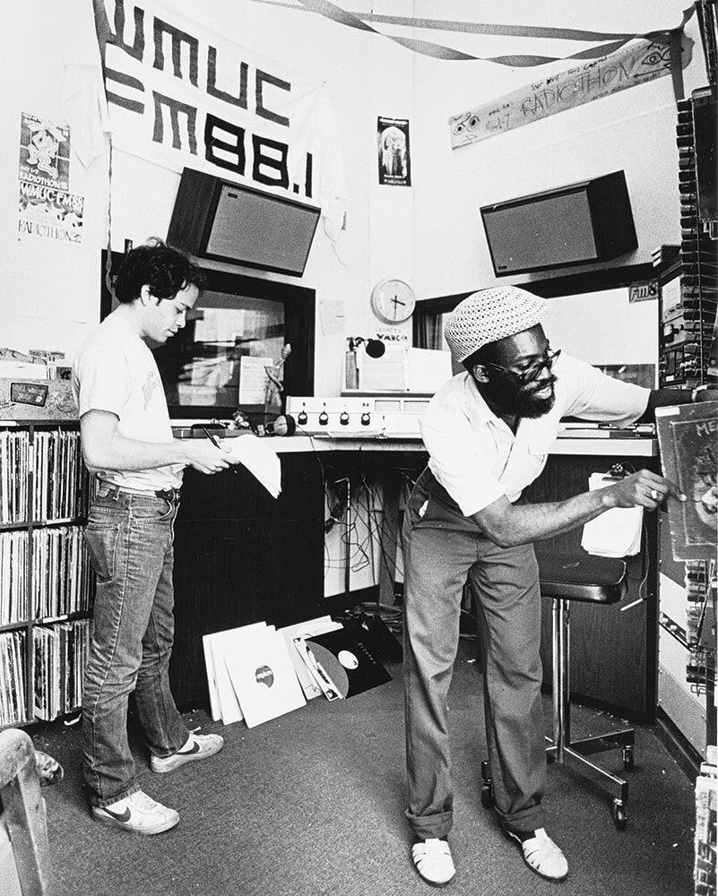 two students review tally sheets with WMUC FM88.1 on the wall behind them