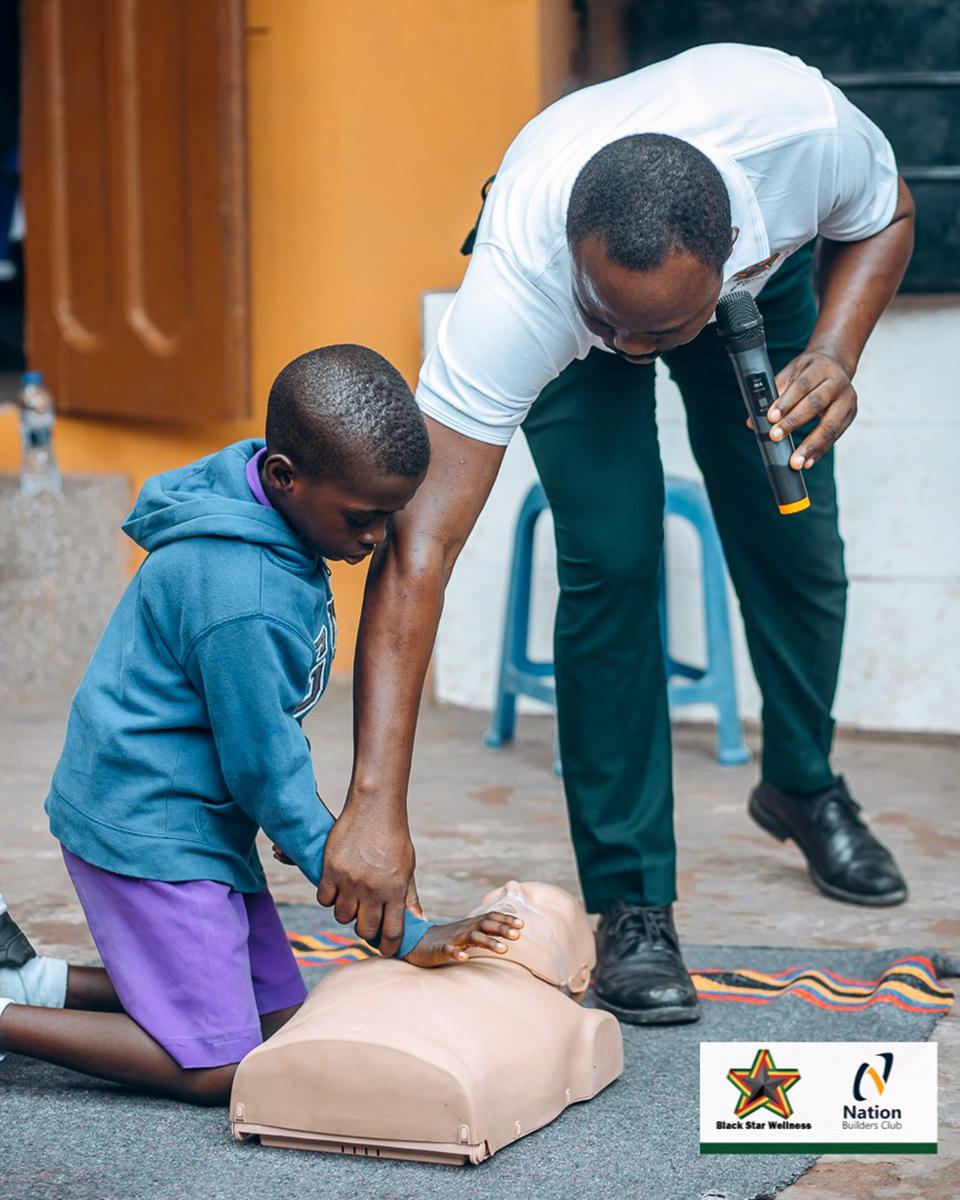 A man holding a microphone helps a youth learn CPR on a dummy