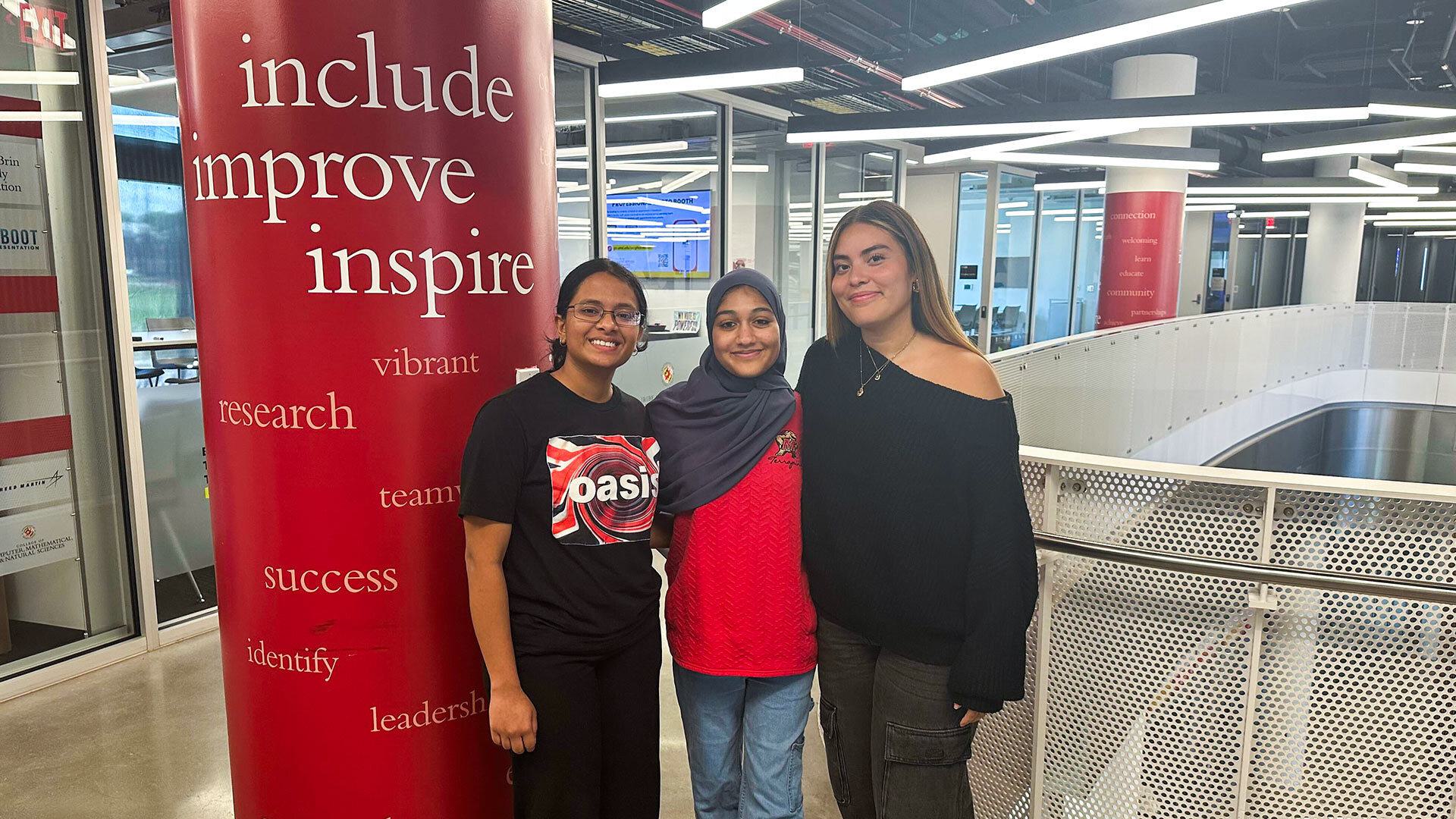 three women pose in front of pillar that reads, "include, improve, inspire"