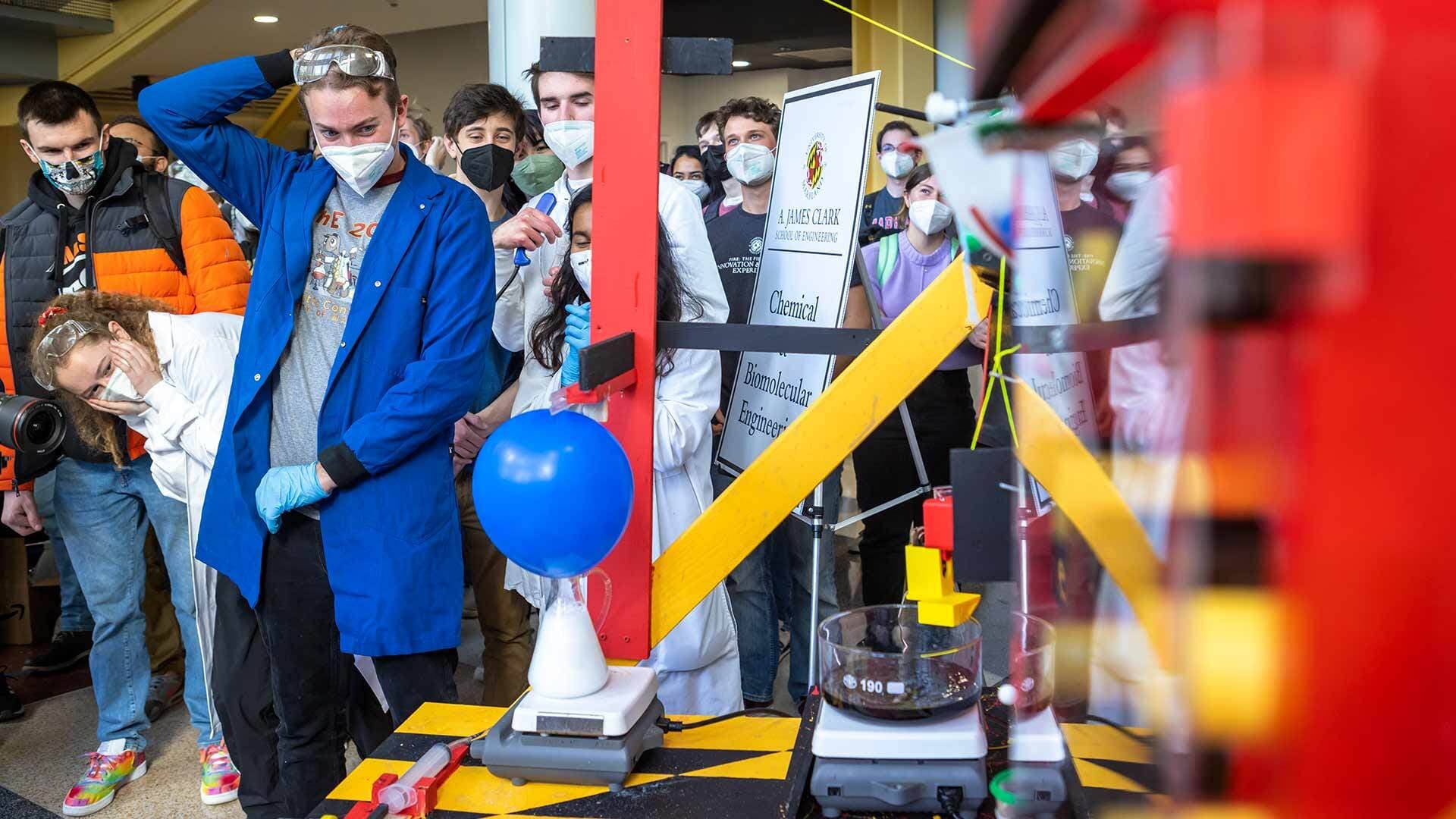 A student in a blue lab coat, protective glasses on her head and face mask, surrounded by other students in masks, watches a blue balloon on top of a beaker filled with a white substance