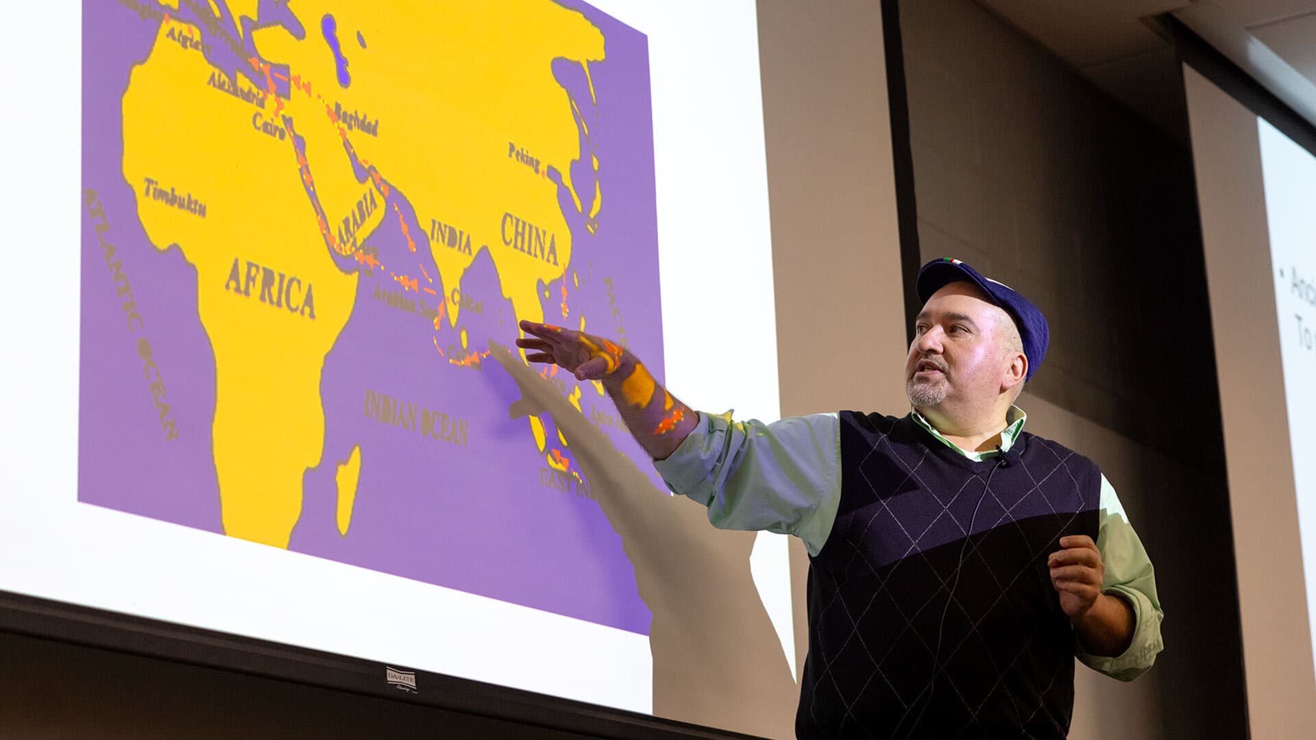 A professor teaches in front of a screen with a world map
