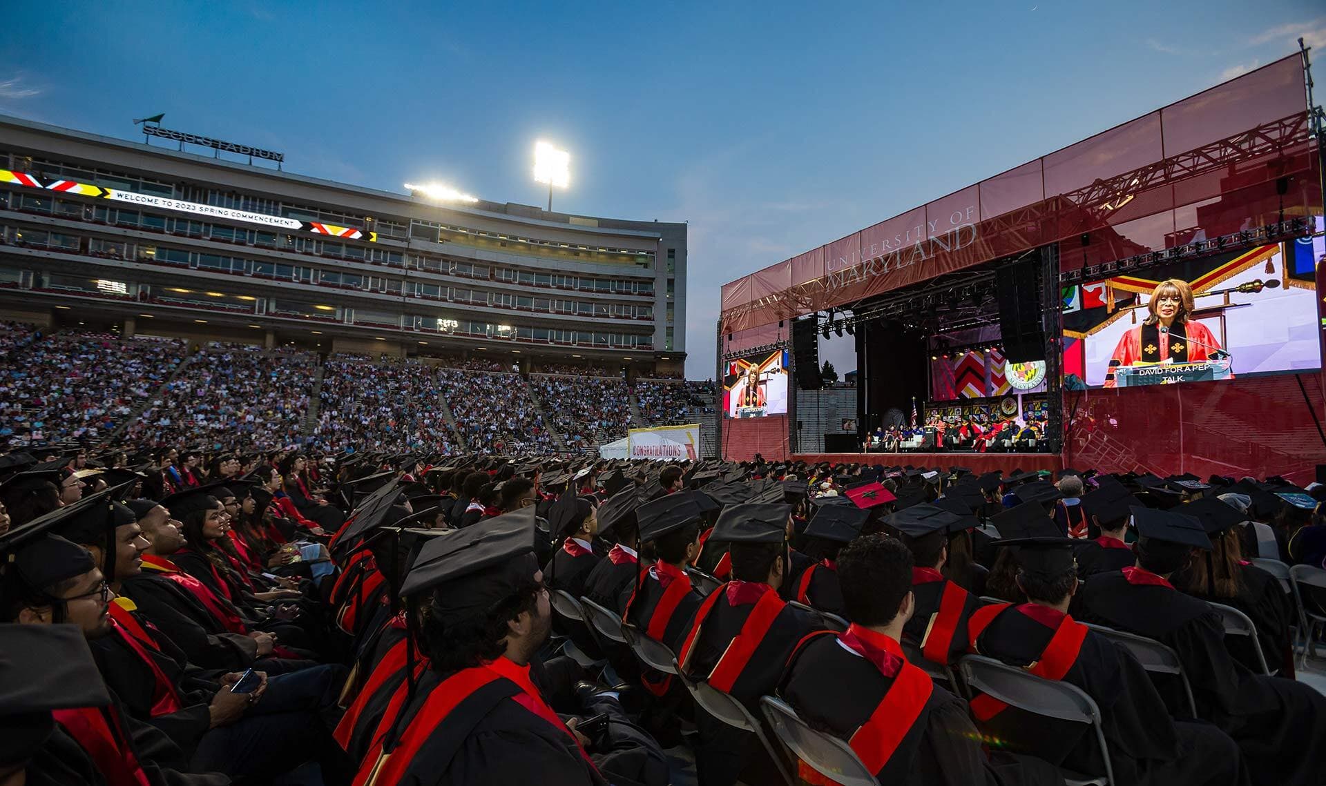 Students in caps and gowns sit in a stadium listening to a Gayle King speak on stage