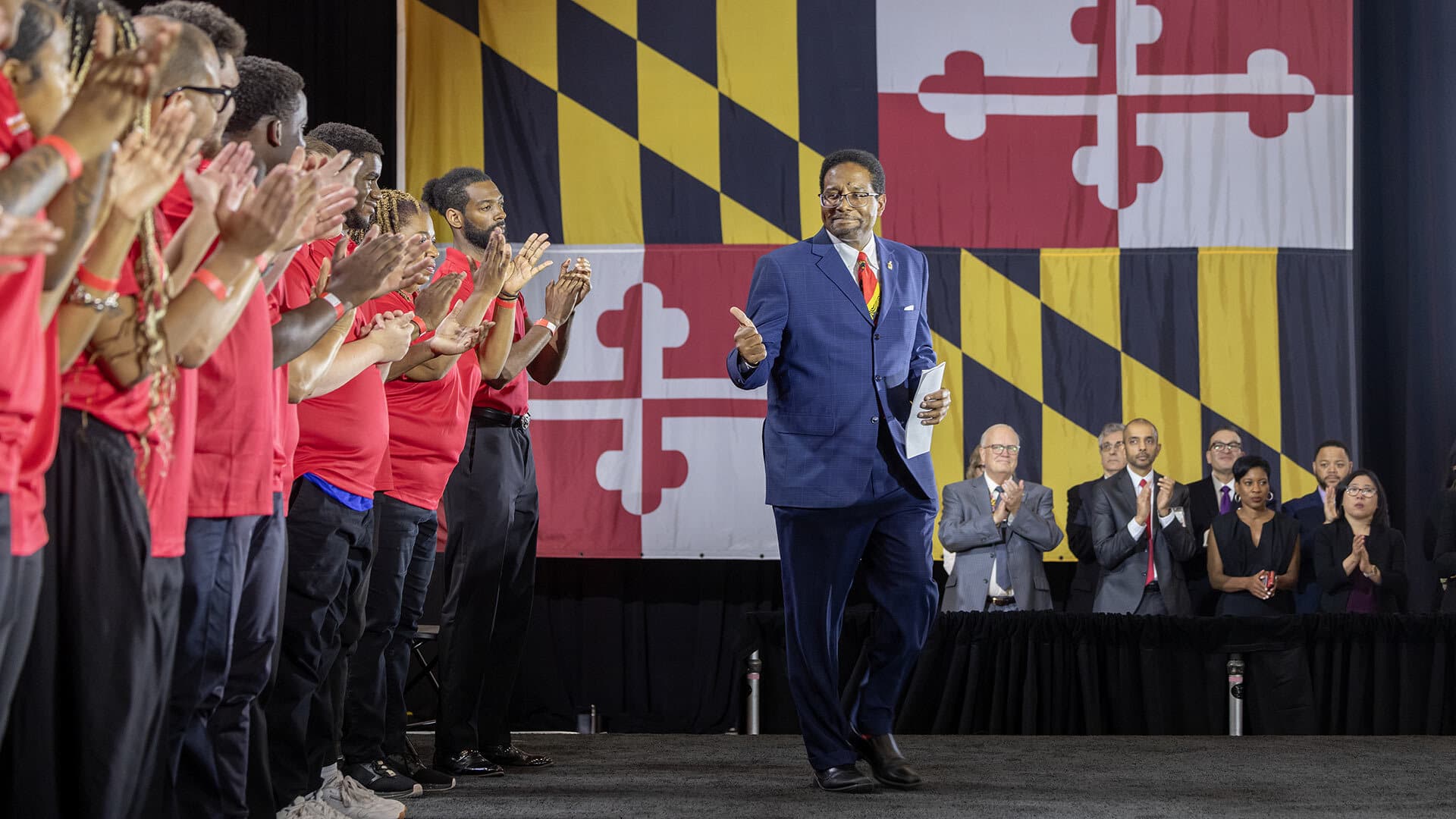 man on stage in front of giant Maryland flag gives thumbs-up sign to a line young people