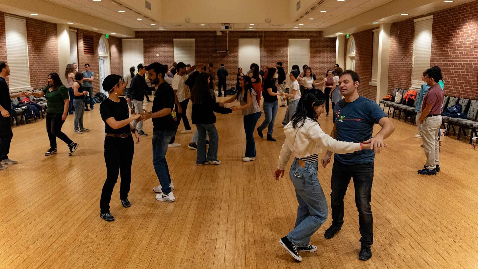 Numerous couples dance in a ball room with a hardwood floor
