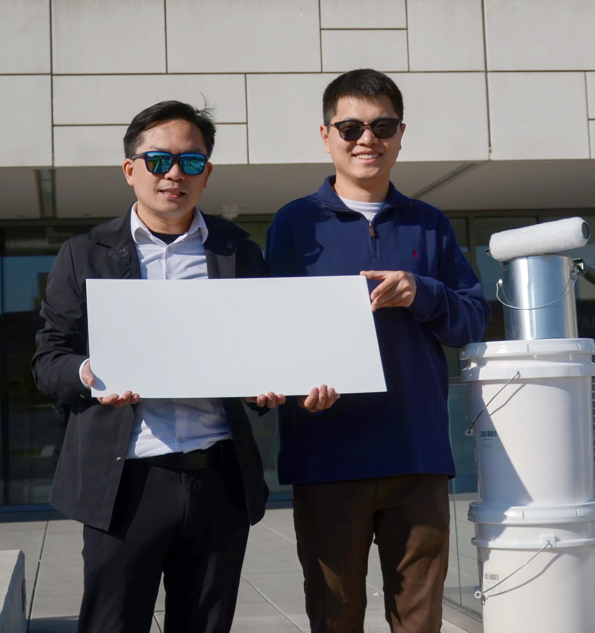 two men  hold a white rectangular piece of metal in front of a white building