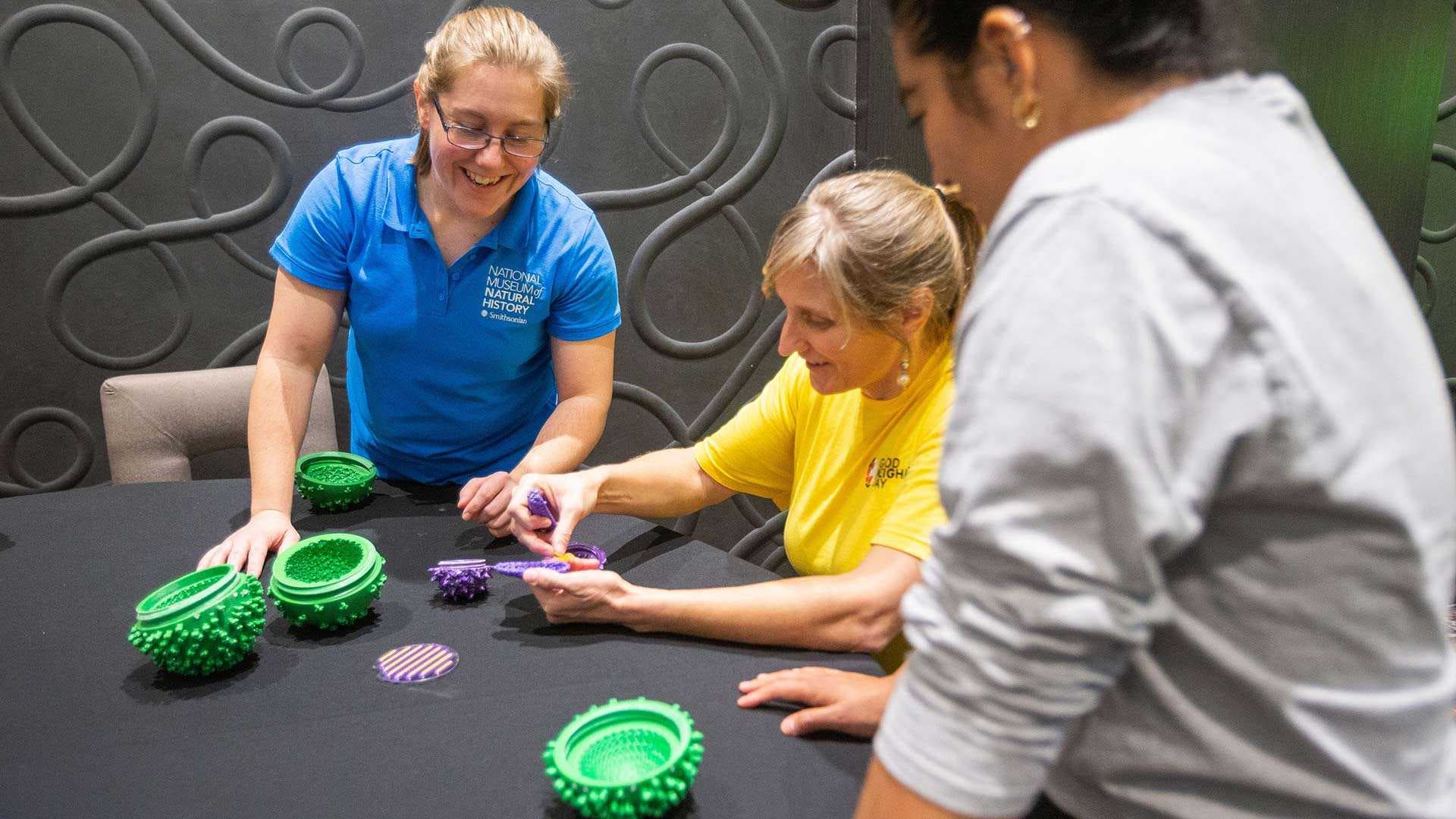 three women look at 3d printed virus models at a table
