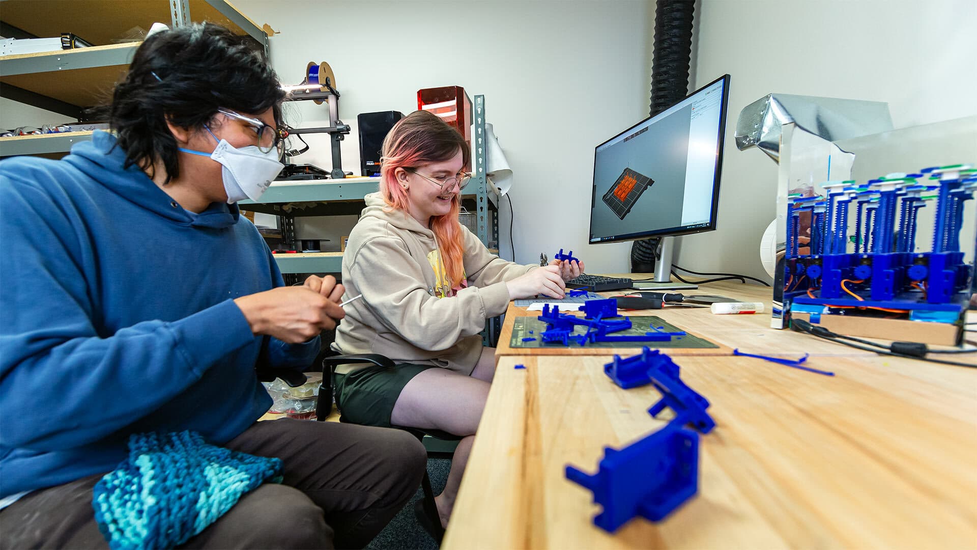 two students sit at a desk and work on blue 3D-printed objects