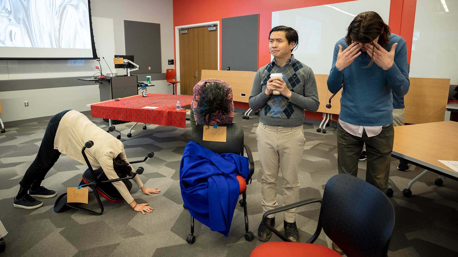one faculty member bends over a chair with a sign that reads, "forced." Another leans with head down on chair with sign that reads, "confused." A third holds a coffee cup, while a fourth covers their eyes with their hands
