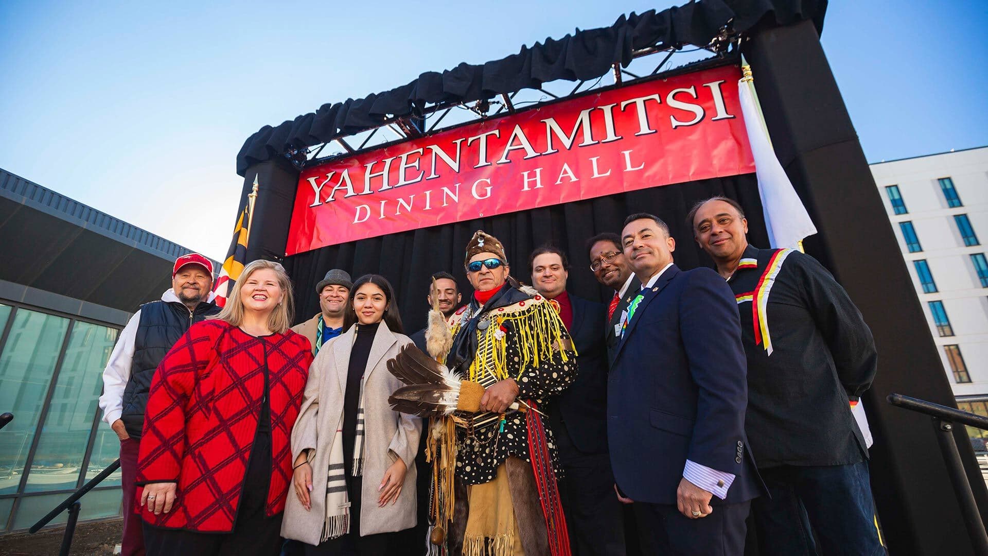 A crowd of people stand under a red banner that says "Yahentamitsi Dining Hall"