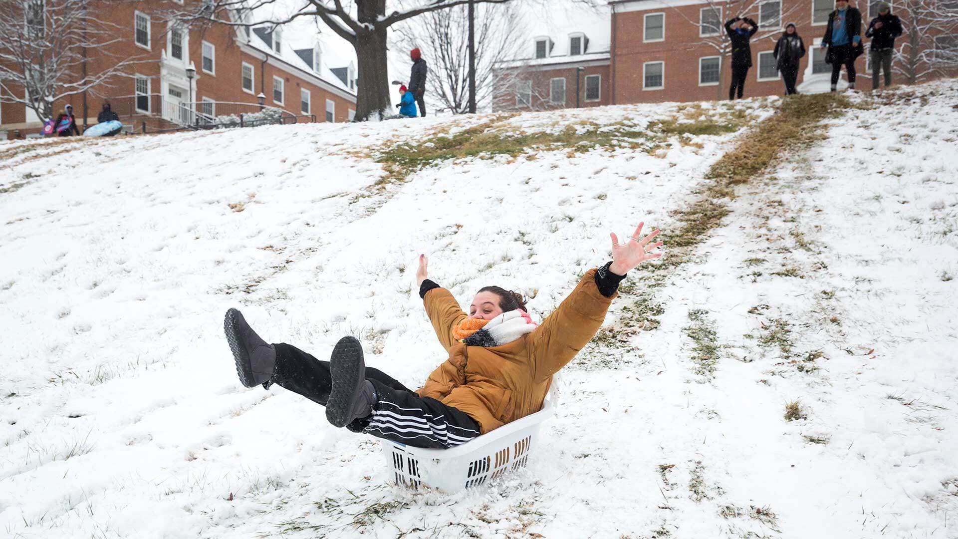 Student sleds down snowy hill in laundry basket
