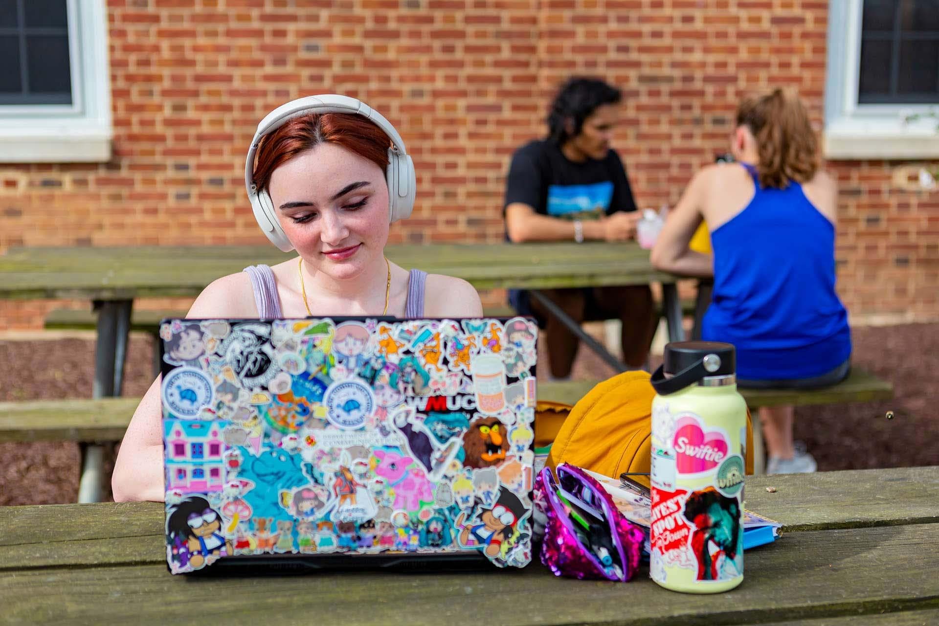 student wears white headphones and works on laptop covered with stickers while sitting at picnic table